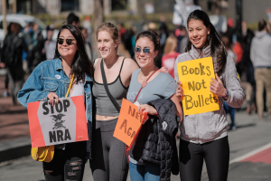 Vier Frauen marschieren auf einer Straße, jede hält ein Schild mit der Aufschrift "Stop NRA", mit einem unscharfen Hintergrund von Menschen, Fahrzeugen, Pfählen, Bäumen und Gebäuden.