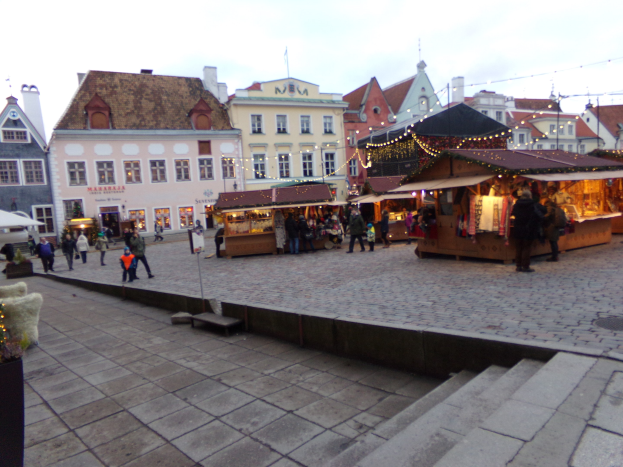 Ein belebter Weihnachtsmarkt in Tallinn, Estland mit Menschen um geschmützte Stände, festlich geschmütkte Gebäude, bewölktem Himmel, Treppen und Topfpflanzen.