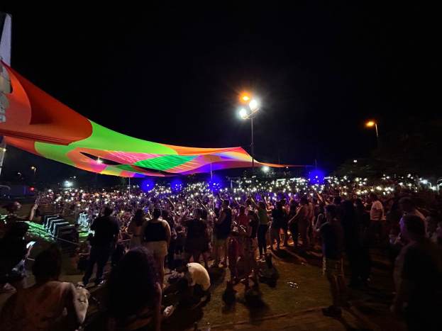 Eine große Menschenmenge steht nachts vor einem Zelt, das von bunten Lichtern beleuchtet ist und von beleuchteten Pfählen umgeben ist, mit einem Gebäude links und dem Nachthimmel im Hintergrund.