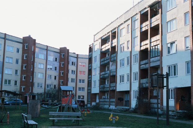 Wohngebäude mit Spielplatz davor, umgeben von Gras, Bänken, Schaukeln, Fahrzeugen, Bäumen und einem klaren blauen Himmel.