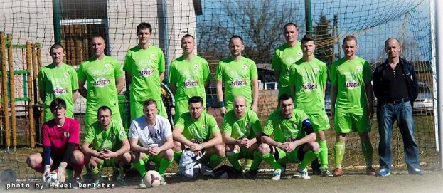 Gruppe von Männern in grünen T-Shirts auf einem Fütbolfeld mit einem Ball und einem Tor, umgeben von Bäumen und Gebäuden.