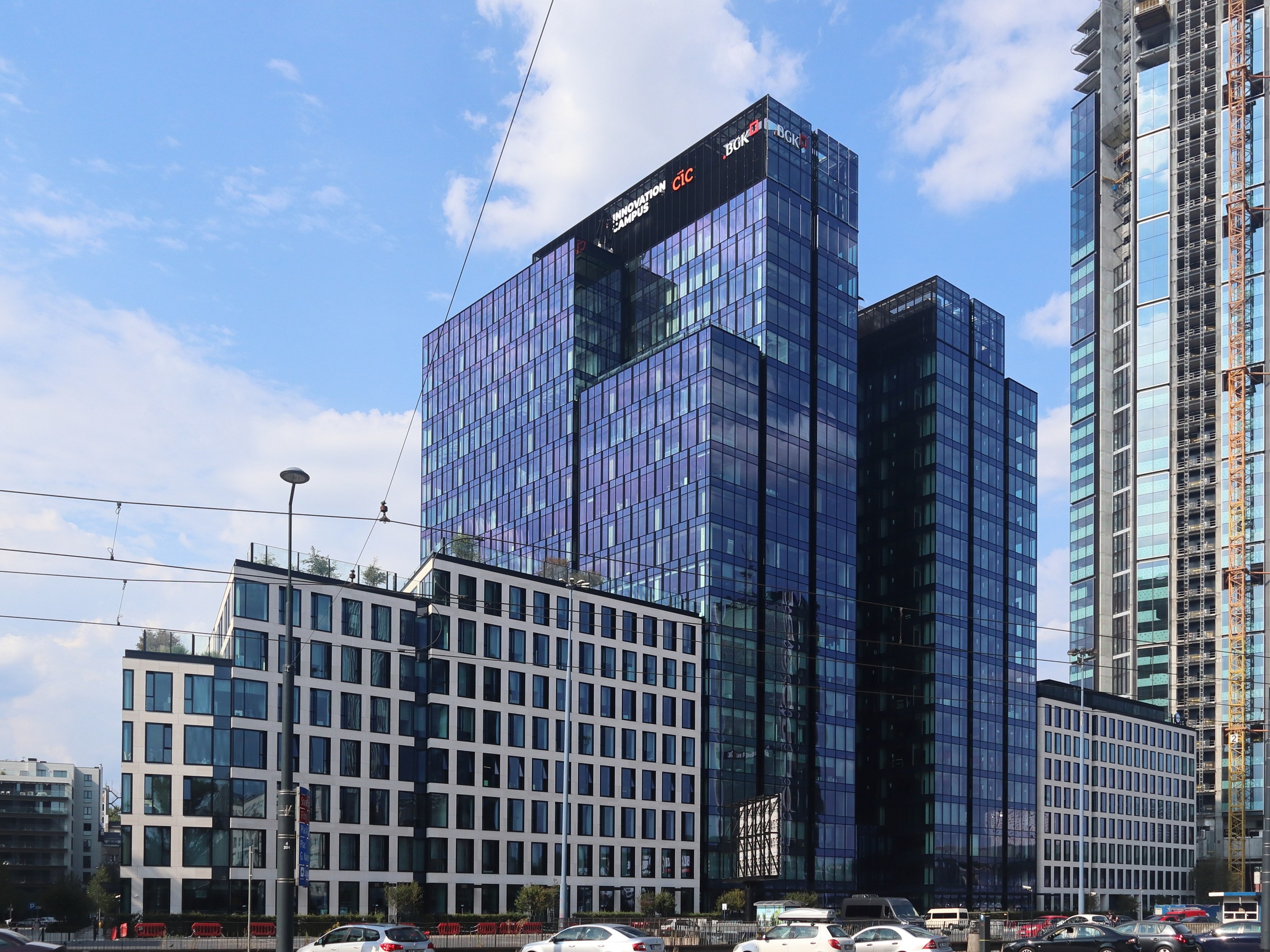 Zentrale der Deutschen Bank in Frankfurt, Deutschland, ein großes mehrfenstriges Gebäude umgeben von städtischer Infrastruktur einschließlich Straßenlaternen, Fahrzeugen und Vegetation unter einem bewölkten Himmel.