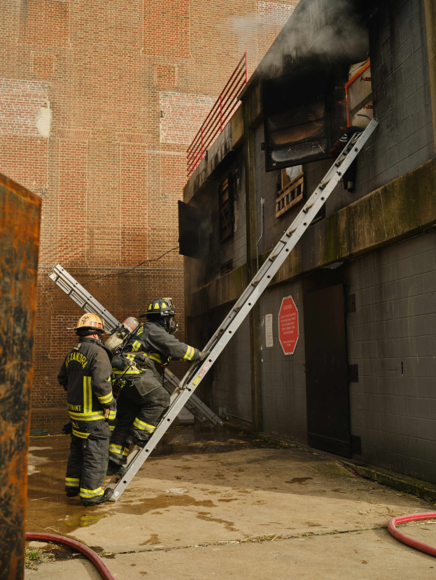 Feuerwehrleute mit Helmen klettern eine Leiter zu einem rauchgefüllten Gebäude hinauf, mit einem Metallobjekt links und Rohren auf dem Boden.