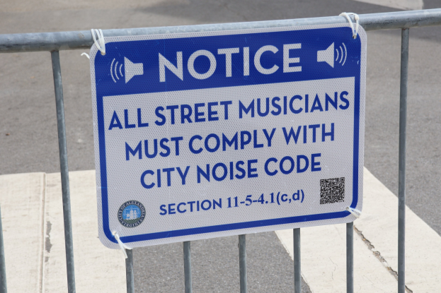 Anschlagtafel an einem Zaun mit einem Schild, das Straßenmusikanten zur Einhaltung des städtischen Lärmschutzes auffordert, mit einer Straße im Hintergrund.