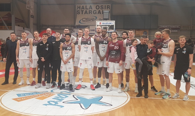 Gruppe von Männern und Kindern auf einem Basketballfeld mit Schildern, die "Hala Osir Starga" lesen, sitzende Zuschauer, ein Basketballkorb, Metallstangen, Deckenlampen und eine Wand im Hintergrund.