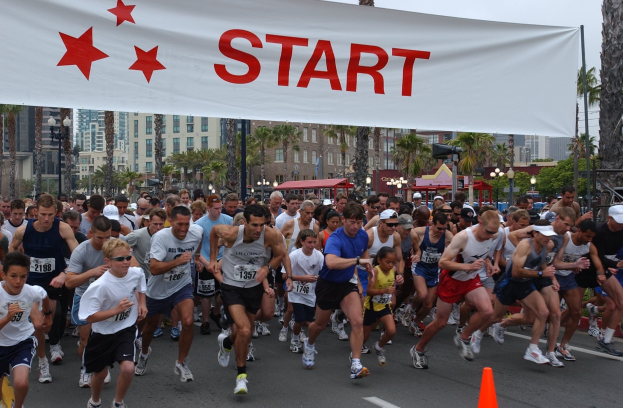 Gruppe von Läufern bei einem Marathon an einer Verkehrskegel vorbei, mit einem Banner und Gebäuden im Hintergrund unter einem klaren blauen Himmel.