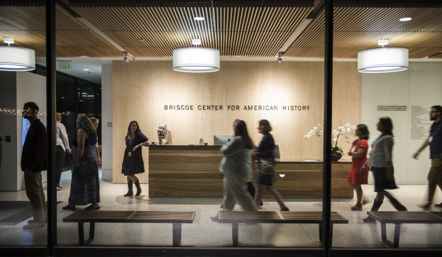 Innenansicht einer Lobby im Briscoe Center for American History, die Menschen beim Gehen, Glastüren, Bänke, einen Tisch mit Gegenständen, einen Topf mit einer Pflanze, Wandtext, ein Schild und Deckenleuchten zeigt.