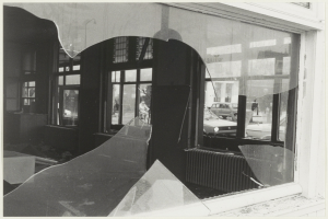 Ein Schwarz-Weiß-Foto eines zerbrochenen Fensters in einem Gebäude, mit Glasscherben drumherum, das Fahrzeuge, Gebäude und Bäume im Hintergrund zeigt.