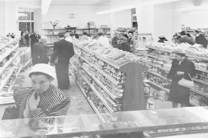 Schwarzes und weißes Foto von Menschen beim Einkaufen in einem Supermarkt, die Taschen halten, mit Lebensmittelregalen, einer Uhr an der Wand und Deckenbeleuchtung.
