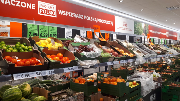 Ein Geschäft mit Kartons voller verschiedener Früchte und Gemüse, jeweils mit Preisschildern, Text- und Bildtafeln an den Wänden und Deckenbeleuchtung.