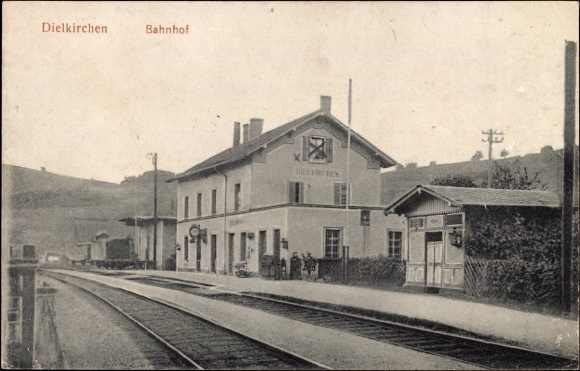 Schwarzes und weißes Foto einer Bahnstation in Dielkirchen, Deutschland, mit einem Zug auf den Schienen, umgeben von Häusern, Strommasten, Bäumen, Menschen und Bergen sowie einem klaren Himmel im Hintergrund.