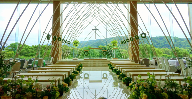 Eine Trauungszeremonie in einem großen Saal mit halbrunder Bestuhlung, geschmückt mit Blumen und Grünpflanzen, vor einer Kulisse aus Bäumen, Hügeln und einem klaren Himmel.