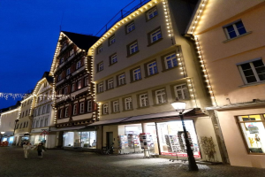 Headsteinstraße in Rothenburg ob der Tauber, Deutschland, geschmückt mit Weihnachtslichtern, mit Menschen und einem geparkten Fahrrad, flankiert von Gebäuden mit Fenstern unter einem sichtbaren Himmel.