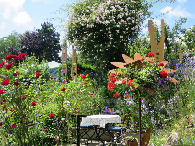 Ein farbenfroher Garten auf dem Chelsea Flower Show mit einem Tisch und Stühlen in der Mitte, bedeckt von einem Tuch, umgeben von Blumen, Pflanzen, Bäumen und einem bewölkten Himmel.