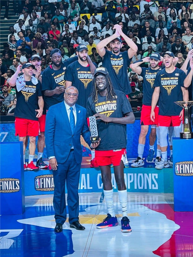 Eine Gruppe von Männern auf einem Basketballfeld feiert mit Trophäen, von denen zwei im Hintergrund auf Podesten stehen, während andere zuschauen.