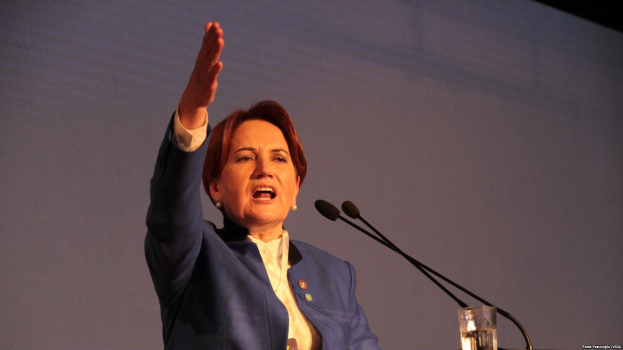 Frau steht an einem Rednerpult mit einem Bildschirm dahinter und hält eine Rede, während sie ein Glas Wasser in der Hand hält, mit zwei Mikrofonen vor ihr.