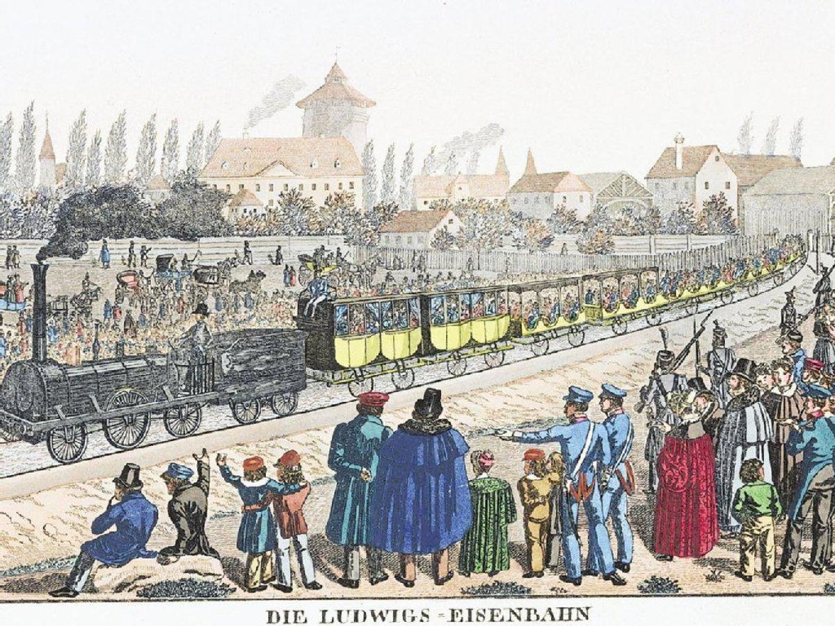 Eine Gruppe von Menschen, die um einen Zug auf einem Gleis stehen, mit einigen im Zug und anderen außerhalb, in der Nähe von Gebäuden und Bäumen, mit dem Text "Ludwigs-Eisenbahn, der erste Bahnhof in Deutschland" unten.