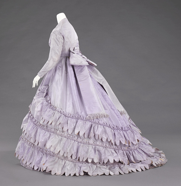 Eine Schaufensterpuppe in einem lila, langärmligen Ballkleid mit engem Oberteil, vollem Rock mit Rüschen und leichtem, luftigem Stoff vor einem grauen Hintergrund.