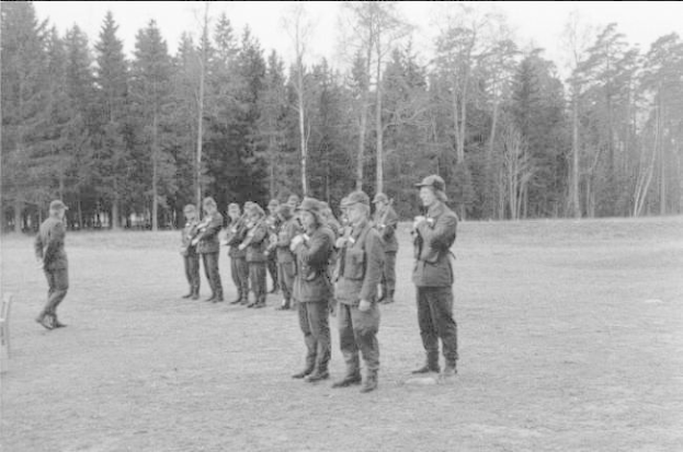 Schwarzes und weißes Bild einer Gruppe von Männern in Militäruniformen mit Mützen und Gewehren, die in einem Feld mit Bäumen und einem klaren Himmel im Hintergrund stehen, wahrscheinlich während eines Trainingsübungs.