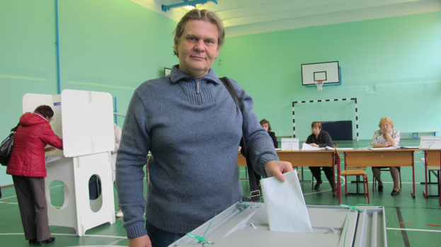 Ein Mann steht vor einer Wahlurne und hält ein Blatt Papier in der Hand, mit Menschen, die hinter ihm sitzen und stehen, und einem Basketballkorb an der Wand im Hintergrund.