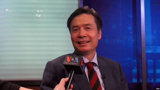 Ein Mann im Anzug mit einem Mikrofon in der Hand, vor einem Fenster und einer Wand stehend.