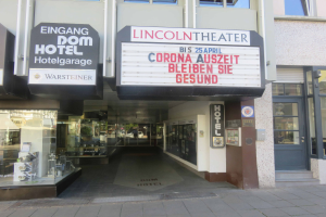 Das Lincoln Theater in Berlin, Deutschland - ein Gebäude mit Glasfenstern und -türen sowie einer Tafel mit Text, die verschiedene Objekte im Inneren zeigt und so einen Eindruck von einer pulsierenden Stadtlandschaft vermittelt.