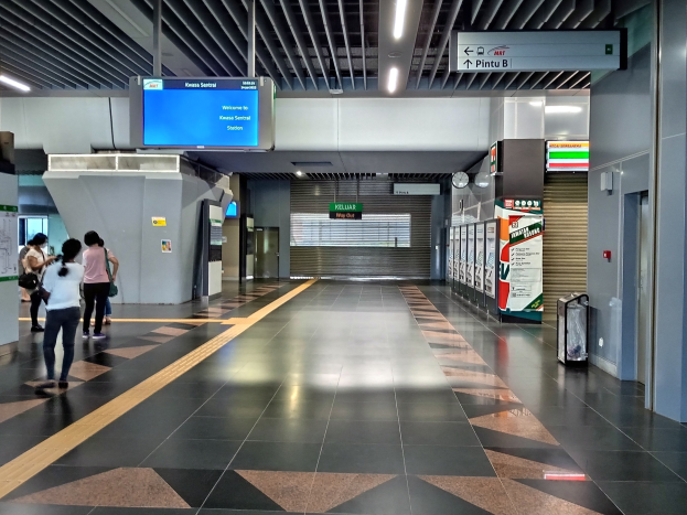 Menschen gehen durch einen Bahnhof mit einem Gepäckwagen auf der rechten Seite, Deckenleuchten, einer Uhr an der Wand und Informationsanzeigen.