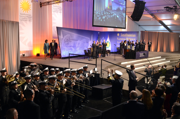 Eine Gruppe von Menschen auf einer Bühne im Smithsonian National Museum of African American History and Culture, einige halten Musikinstrumente oder Kameras, mit Treppen, Bannern, einem Bildschirm und Deckenlampen im Hintergrund.