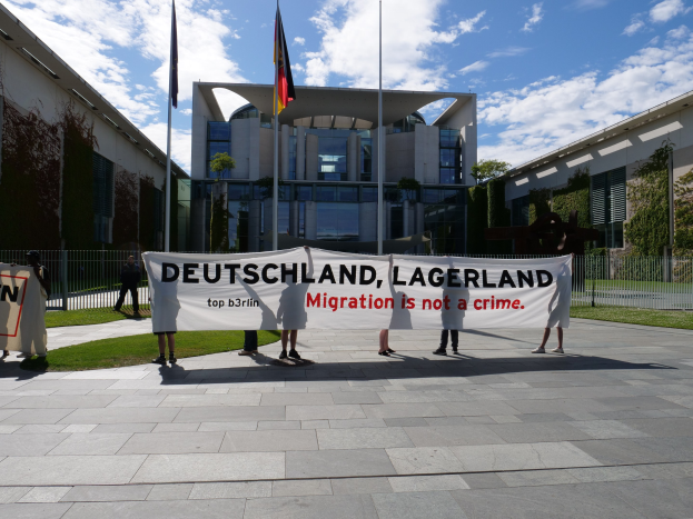 Eine Gruppe von Menschen hält ein Transparent mit der Aufschrift "Deutschland, Lagerland Migration ist kein Verbrechen" vor einem Gebäude mit Fenstern, umgeben von Gras, einem Metallzaun, Pflanzen, Bäumen und Fahnenmasten gegen einen bewölkten Himmel.