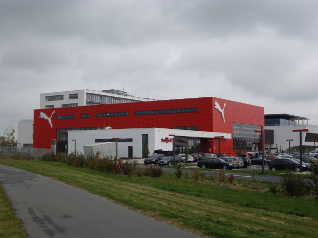 Die Puma-Fabrik in Deutschland, mit Industriegebäuden, Fahrzeugen auf einer Straße, Straßenlaternen, Bäumen, Pflanzen, Gras und einem bewölkten Himmel im Hintergrund.