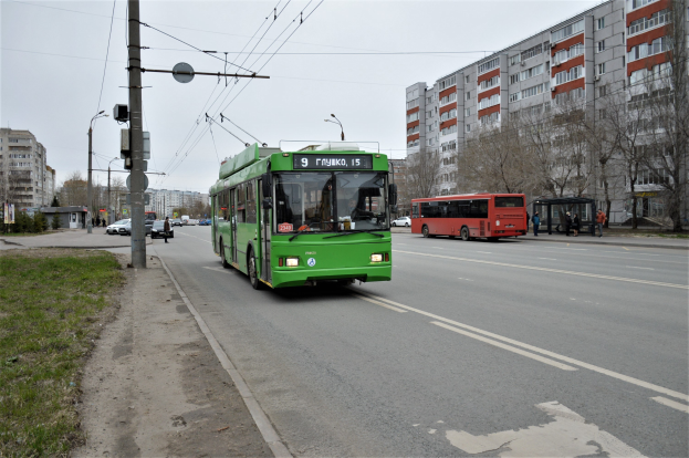 Ein grünter Bus fährt auf einer Straße mit hohen Gebäuden, Gras auf der linken Seite, Strommasten mit Drähten, Bäumen im Hintergrund und einem klaren blauen Himmel.