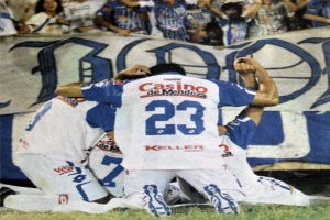 Eine Gruppe von Fußballspielern, die ein Tor auf dem Boden vor einer jubelnden Menge feiern, mit einem Banner mit Text im Hintergrund.