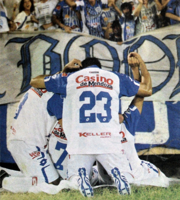 Eine Gruppe von Fußballspielern, die ein Tor auf dem Boden vor einer jubelnden Menge feiern, mit einem Banner mit Text im Hintergrund.