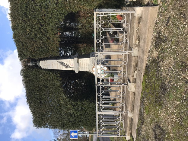 Ein Denkmal für gefallene Soldaten steht in einem Park, umgeben von einem Metallzaun, mit Bäumen im Hintergrund, einer Straße unten und einem bewölkten Himmel darüber.