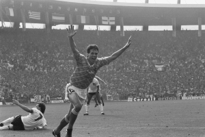 Ein Schwarz-Weiß-Foto eines Fußballspielers, der ein Tor feiert, umgeben von Menschen auf dem Boden, mit Schildern, Fahnen und Pfosten im Hintergrund unter einem klaren Himmel.