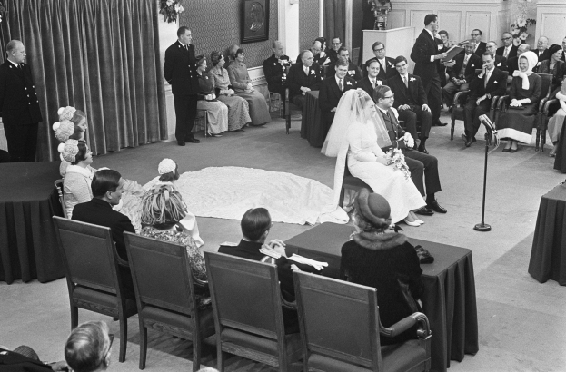 Schwarzes und weißes Foto einer Hochzeitszeremonie mit sitzenden und stehenden Menschen, einem Mann mit Mikrofon, einer Frau mit einem Blumenstrauß, einem Podium mit Mikrofon, Blumenschmuck, einem Vorhang, einem gerahmten Foto an der Wand und einer Tür im Hintergrund.
