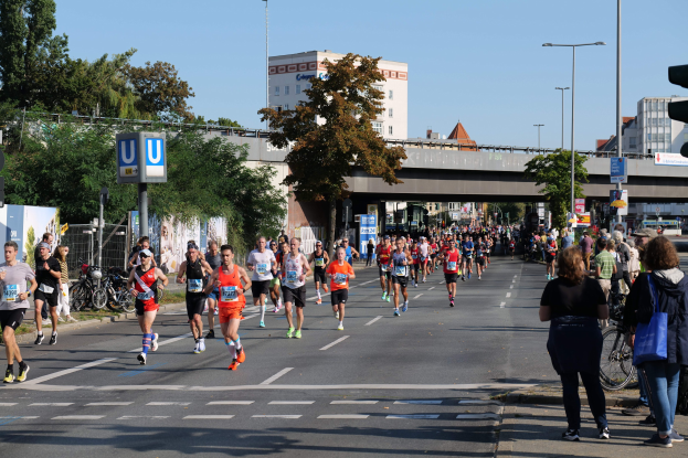 Gruppe von Menschen, die bei einem Marathon auf einer baumgesäumten Straße laufen, mit Laternen, Schildern, Fahrrädern, einem Zaun, Gras, einer Brücke, Gebäuden und einem klaren blauen Himmel.