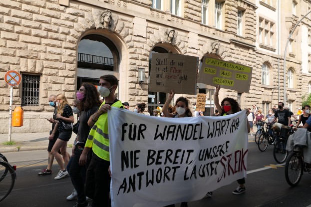 Eine Gruppe von Menschen nimmt an einer Anti-Kriegsdemo in Berlin teil, einige halten Schilder und andere fahren Fahrräder, vorbei an einem Gebäude mit architektonischen Details und Bäumen.