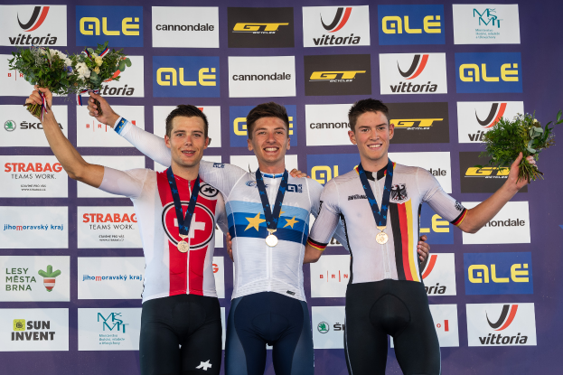 Drei Männer stehen auf einem Podium, jeder mit einer Medaille und einem Blumenstrauß in der Hand, vor einem "Giro d'Italia"-Schild im Hintergrund.