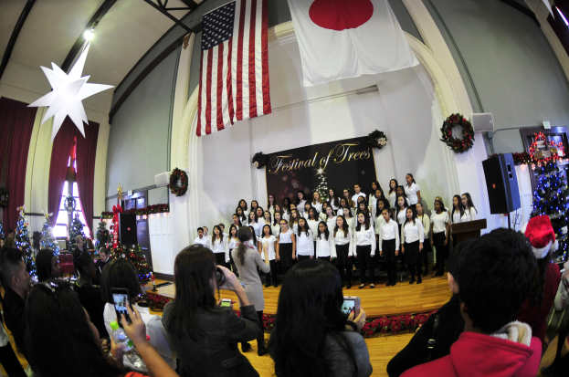 Eine Gruppe von Menschen auf einer Bühne mit Weihnachtsdekoration, einem Podium, Lautsprechern, einer Texttafel und einem Publikum, das Kameras und Handys hält.