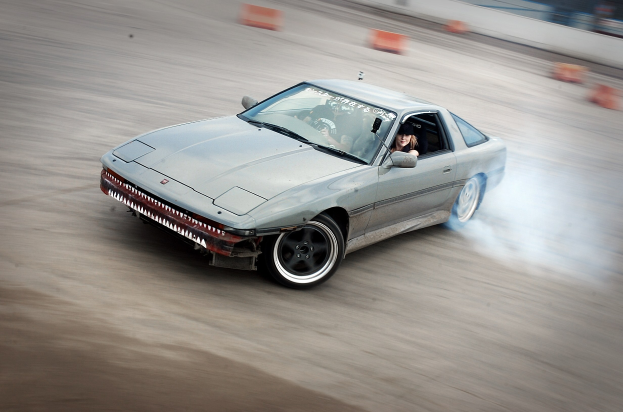 Eine Person in einem Helm fährt ein Auto auf einer Rennstrecke, wobei Rauch austritt, umgeben von orangefarbenen Objekten, mit einem unscharfen Hintergrund, der hohe Geschwindigkeit andeutet.