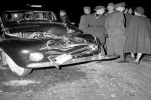 Eine Schwarz-Weiß-Szene eines beschädigten Autos am Straßenrand, mit einer Gruppe von Menschen drumherum, die das Auto besorgt anschauen.