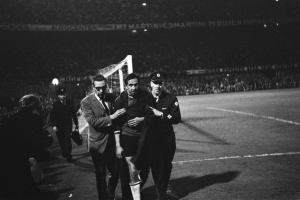 Eine Gruppe von Männern, die auf einem Fußballfeld feiern, mit einem Torpfosten und beleuchteten Rängen im Hintergrund, dargestellt in Schwarz-Weiß.