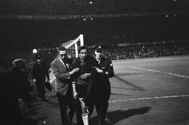 Eine Gruppe von Männern, die auf einem Fußballfeld feiern, mit einem Torpfosten und beleuchteten Rängen im Hintergrund, dargestellt in Schwarz-Weiß.