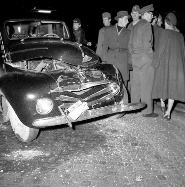 Ein Schwarz-Weiß-Bild eines auf der Straße liegenden Wagens mit eingedellter Motorhaube und zersplitterten Fenstern, umgeben von einer Gruppe besorgter Menschen.