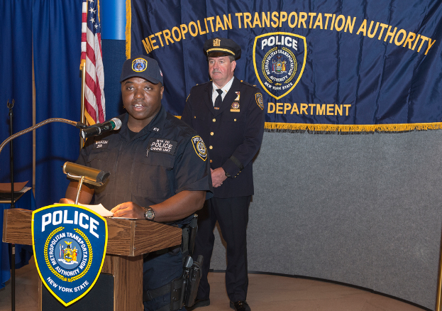 Zwei Polizisten stehen vor einem Rednerpult, einer hält Papiere und spricht in ein Mikrofon, mit einer Fahne, einem Banner und einem Tisch in der Nähe und einer Tafel mit Symbolen und Text dahinter.