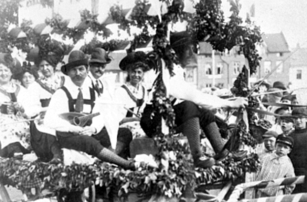 Eine Gruppe von Menschen in traditioneller bayrischer Tracht fährt auf einem mit Laub geschmückten Festwagen mit, einige spielen Musikinstrumente, mit Gebäuden und einem klaren Himmel im Hintergrund, dargestellt in Schwarz-Weiß.