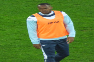 Ein Füßballer in einem orangen T-Shirt und blauen Hosen, der entschlossen über ein saftiges Grünfeld geht.