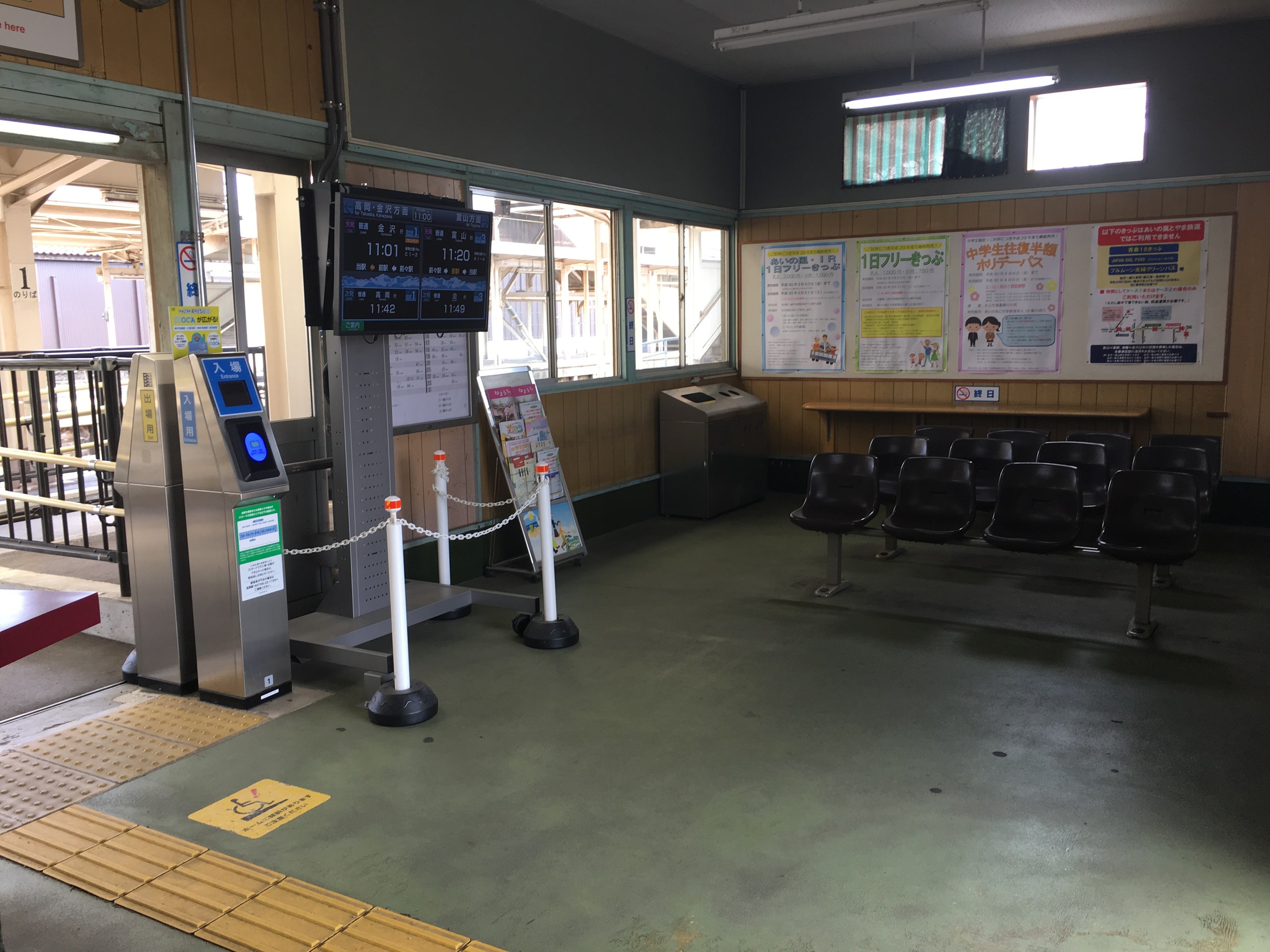 Ein belebter Bahnhofseingang mit Sitzgelegenheiten, einem Service-Schalter, Ticketautomaten, pfostenmontierten Kettenbarrieren, Informationsanzeigen, Wandplakaten, Deckenbeleuchtung und Geländern.