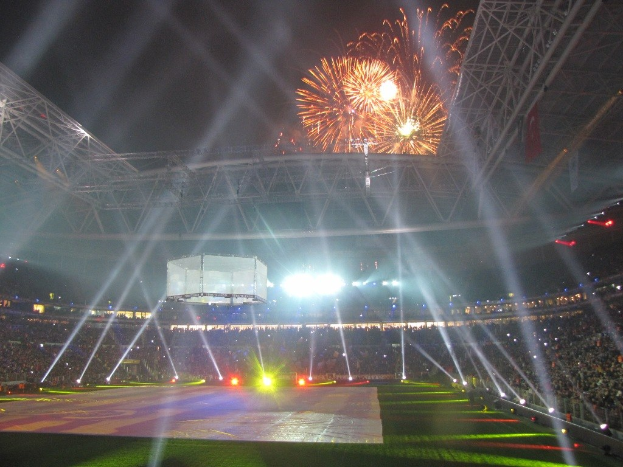 Das Olympische Stadion in London, England, nachts mit hellen Lichtern beleuchtet, umgeben von einer großen Menge, grünem Gras und einem spektakulären Feuerwerk am Himmel.